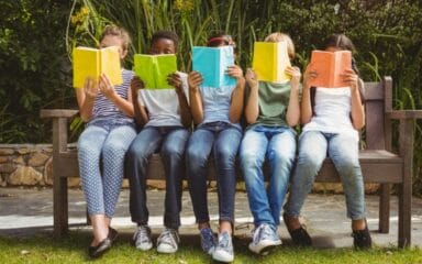 estudiantes sentados en un banco, leyendo, con las caras casi cubiertas por sus libros.