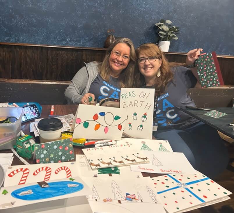 Dos mujeres sonríen ante una mesa cubierta de tarjetas navideñas hechas a mano