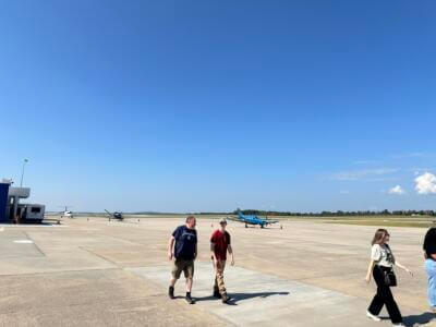 Gente caminando por la soleada pista de un aeropuerto con pequeños aviones de fondo.