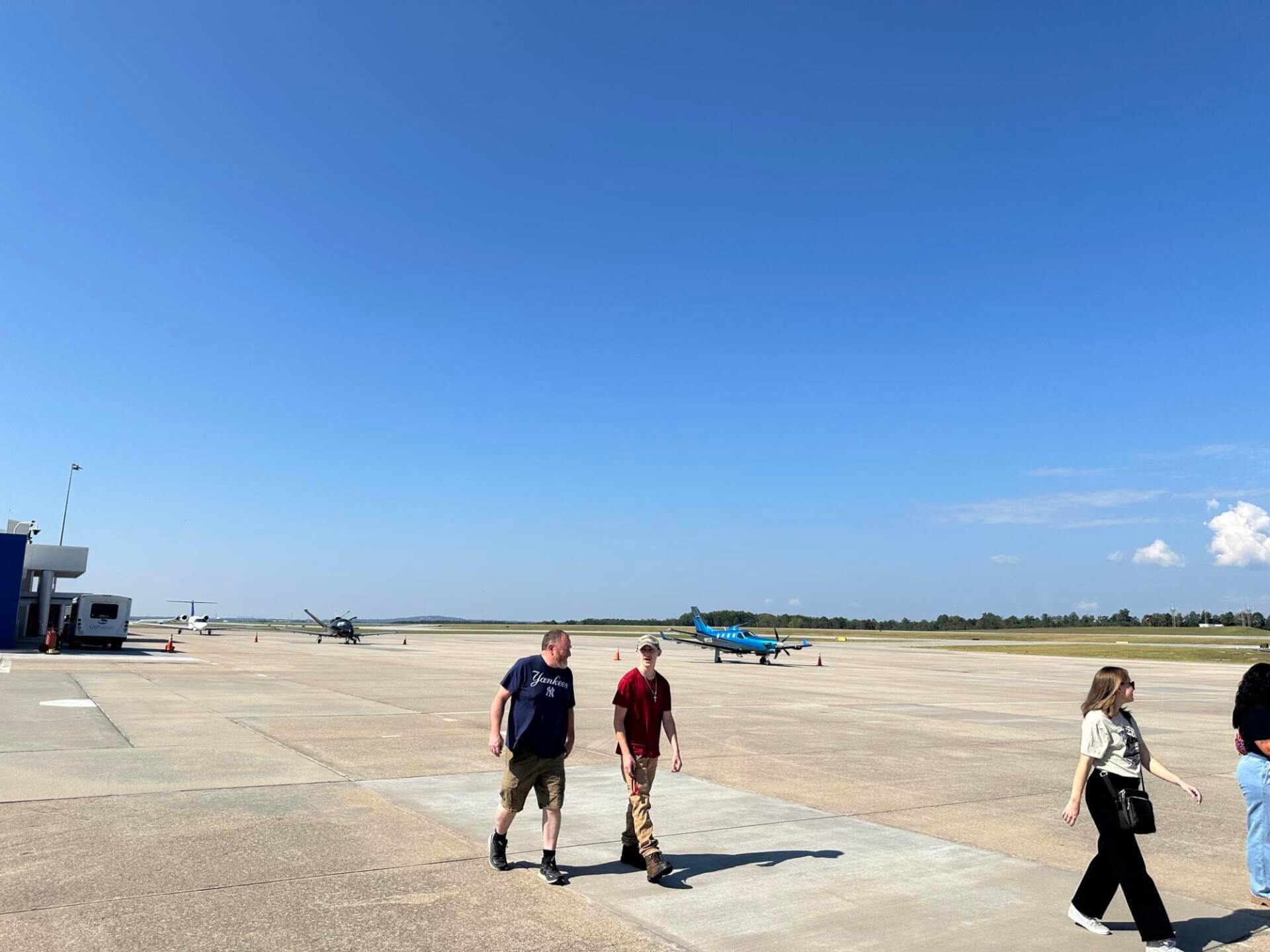 Gente caminando por la soleada pista de un aeropuerto con pequeños aviones de fondo.