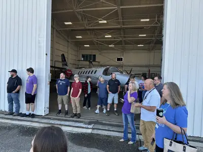 Un grupo de personas se encuentra ante la puerta abierta de un hangar, en cuyo interior se ve parcialmente un pequeño avión.