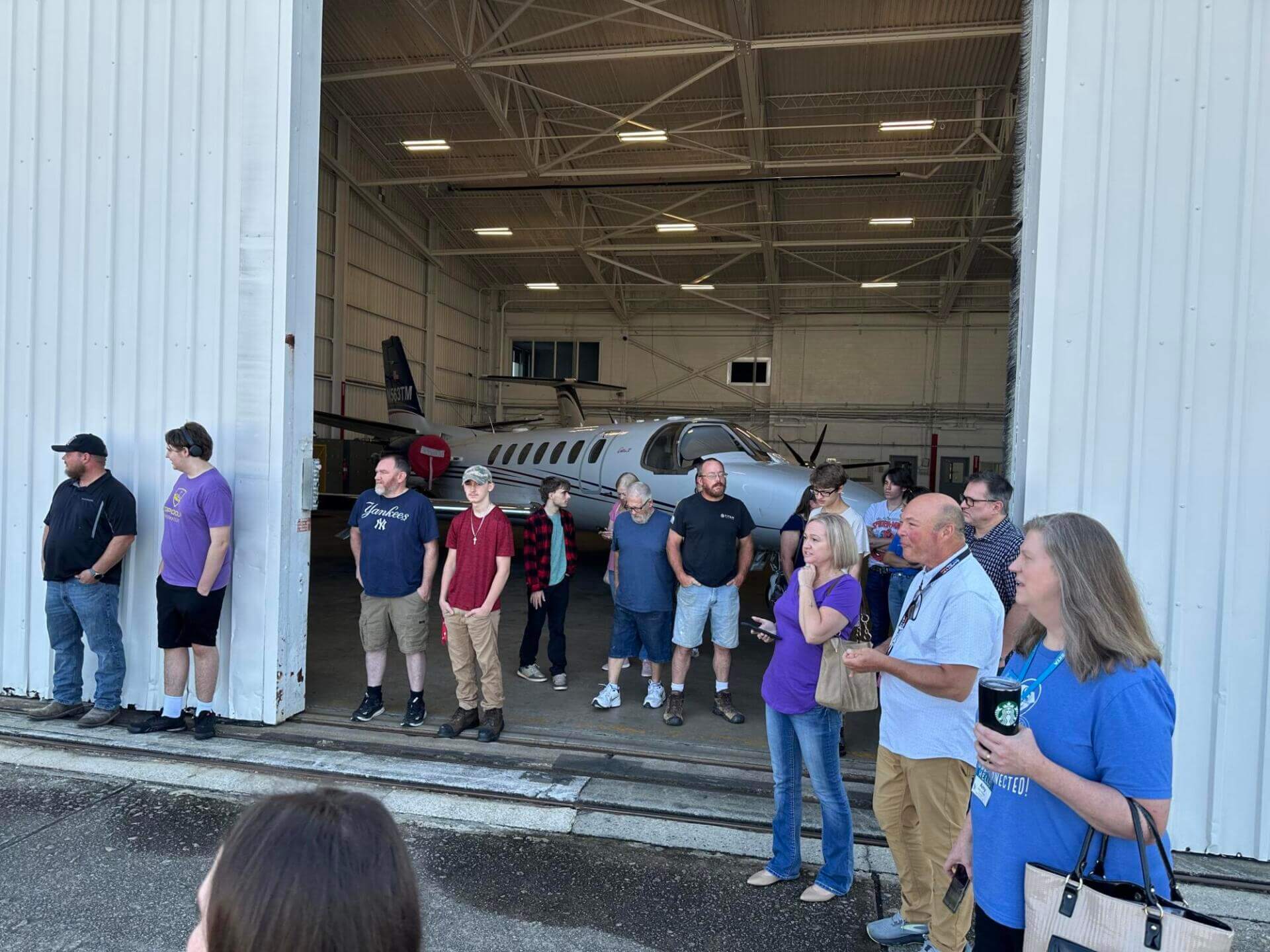 Un grupo de personas se encuentra ante la puerta abierta de un hangar, en cuyo interior se ve parcialmente un pequeño avión.