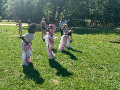 Cuatro niños participan alegremente en una carrera de sacos en un día soleado en un parque cubierto de hierba. Los espectadores observan cómo los niños saltan al unísono.