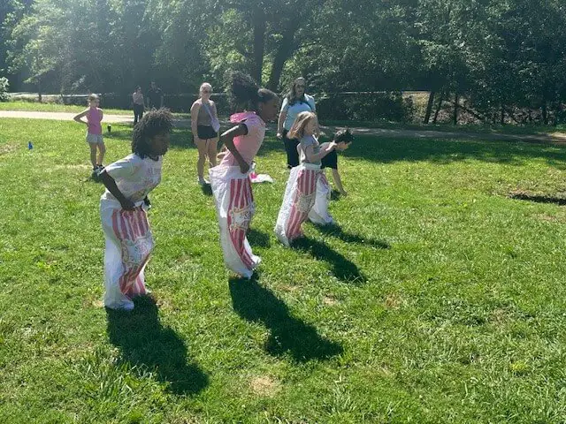 Cuatro niños participan alegremente en una carrera de sacos en un día soleado en un parque cubierto de hierba. Los espectadores observan cómo los niños saltan al unísono.