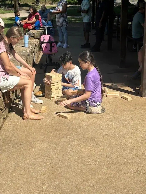 Dos niños juegan con grandes bloques de madera en un camino soleado. Están concentrados y absortos. Cerca, adultos y niños se relajan en un banco de piedra.