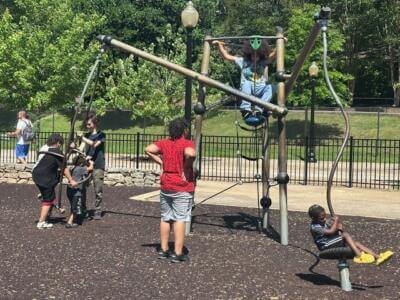 Unos niños juegan en una moderna estructura de un parque. Un niño está sentado en un columpio, mientras otros trepan y se cuelgan de la estructura metálica.