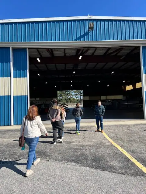 Un grupo de personas camina hacia un gran hangar azul y blanco bajo un cielo azul despejado.