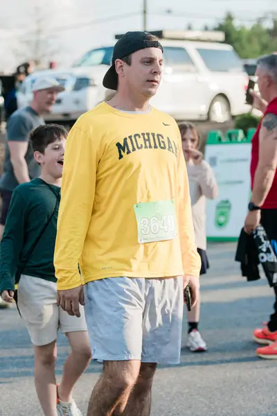 Un hombre con una camiseta amarilla "Michigan" y pantalones cortos grises camina en una carrera, llevando una gorra hacia atrás. Un niño sonriente y otros participantes están en el fondo.