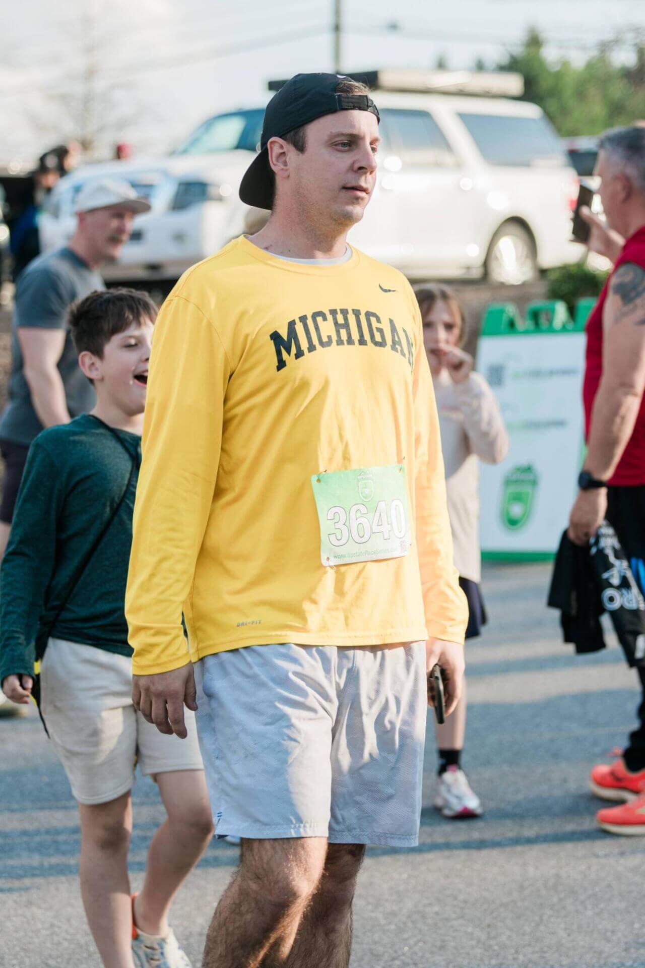 Un hombre con una camiseta amarilla "Michigan" y pantalones cortos grises camina en una carrera, llevando una gorra hacia atrás. Un niño sonriente y otros participantes están en el fondo.