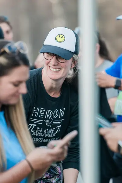 Una persona con gorra sonriente y gafas sonríe mientras se relaciona con los demás en un acto al aire libre. La camiseta "Pi Day Run Walk" indica un ambiente divertido y desenfadado.