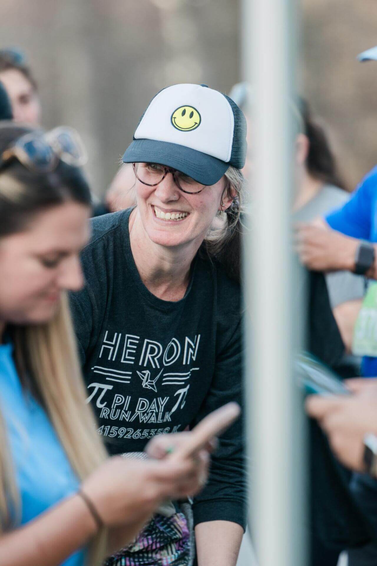 Una persona con gorra sonriente y gafas sonríe mientras se relaciona con los demás en un acto al aire libre. La camiseta "Pi Day Run Walk" indica un ambiente divertido y desenfadado.