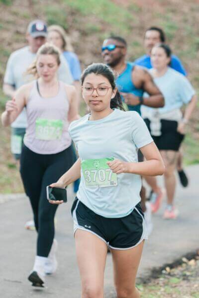 Una mujer joven con ropa deportiva, gafas y un teléfono en la mano, corre en una maratón con otras personas. La escena transmite energía y concentración.