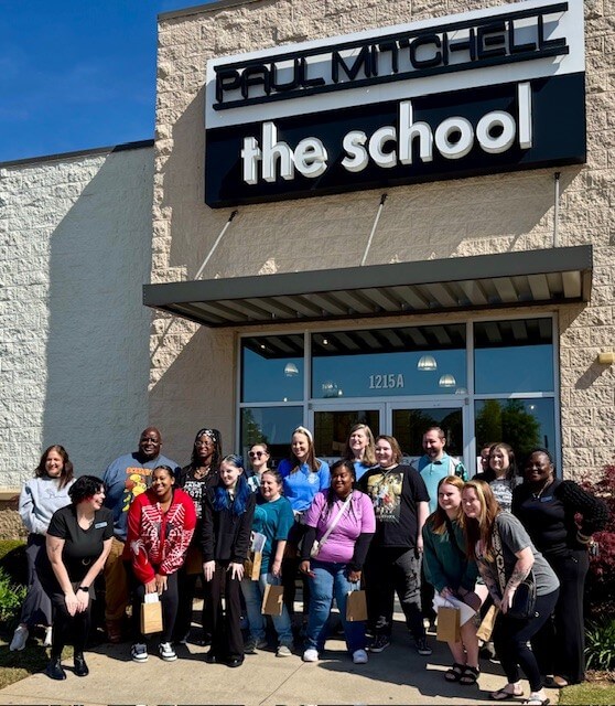 Un grupo heterogéneo de 18 personas sonrientes frente al edificio "Paul Mitchell the School".