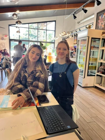 Dos mujeres jóvenes sonríen en el interior de una acogedora cafetería con decoración de madera y luces colgantes.