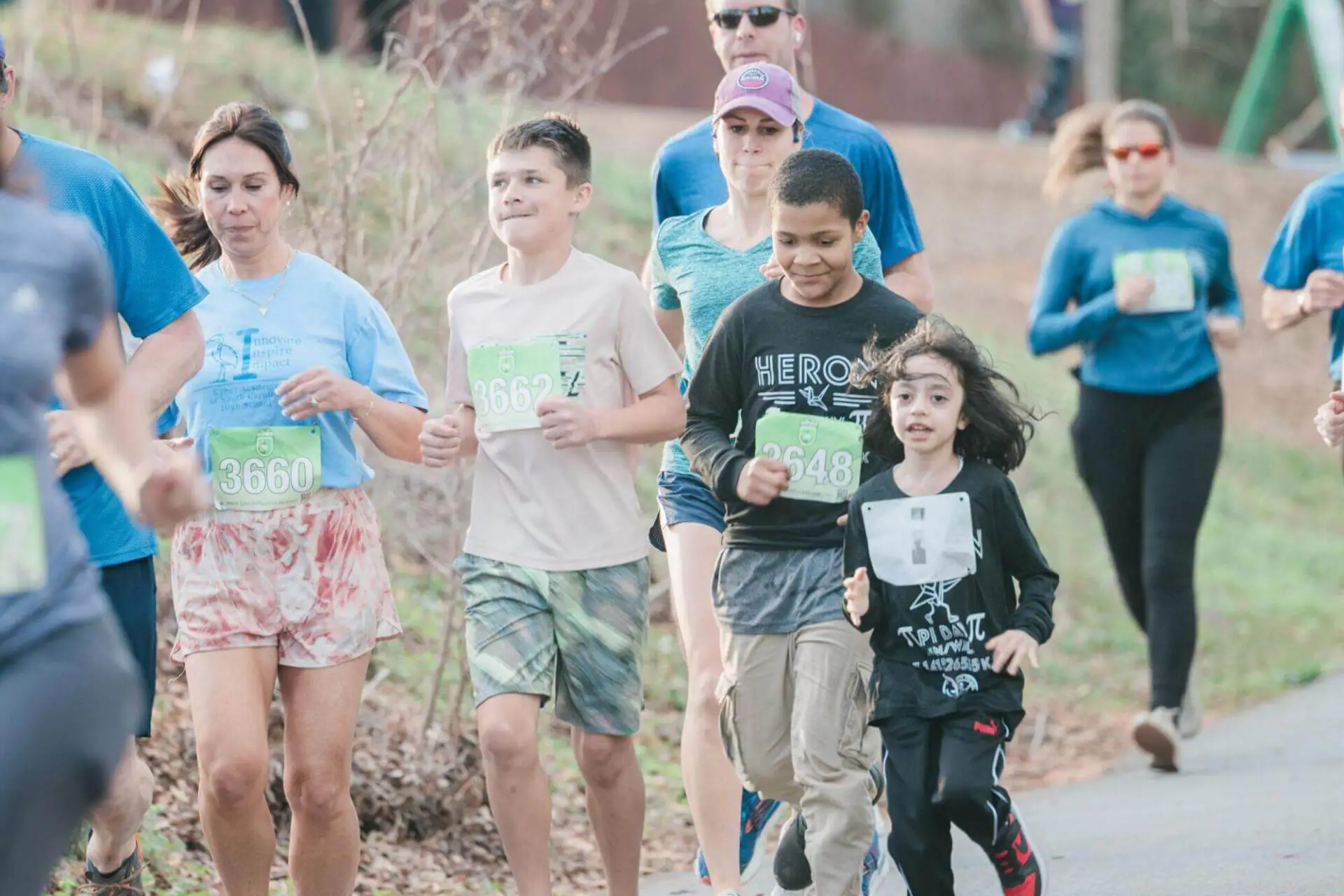 Un grupo diverso de personas, entre las que hay niños y adultos, corren alegremente en una carrera al aire libre llevando dorsales numerados. El ambiente es enérgico y concentrado.