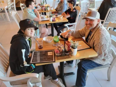 Una persona con sombrero de cubo y otra con gorra sonríen en la mesa de un café con menús.