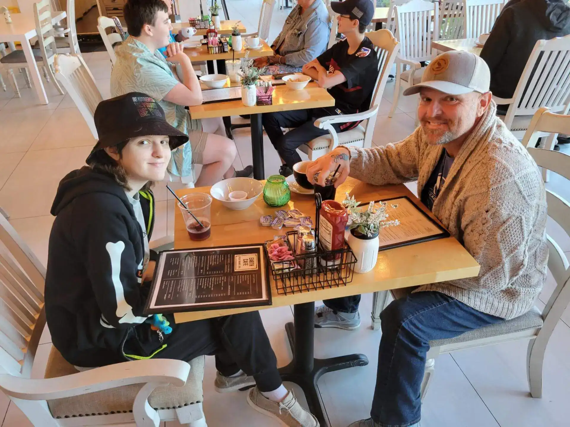 Una persona con sombrero de cubo y otra con gorra sonríen en la mesa de un café con menús.