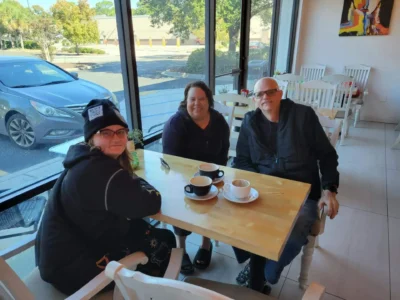 Tres personas se sientan a una mesa de madera en una acogedora cafetería junto a un gran ventanal.