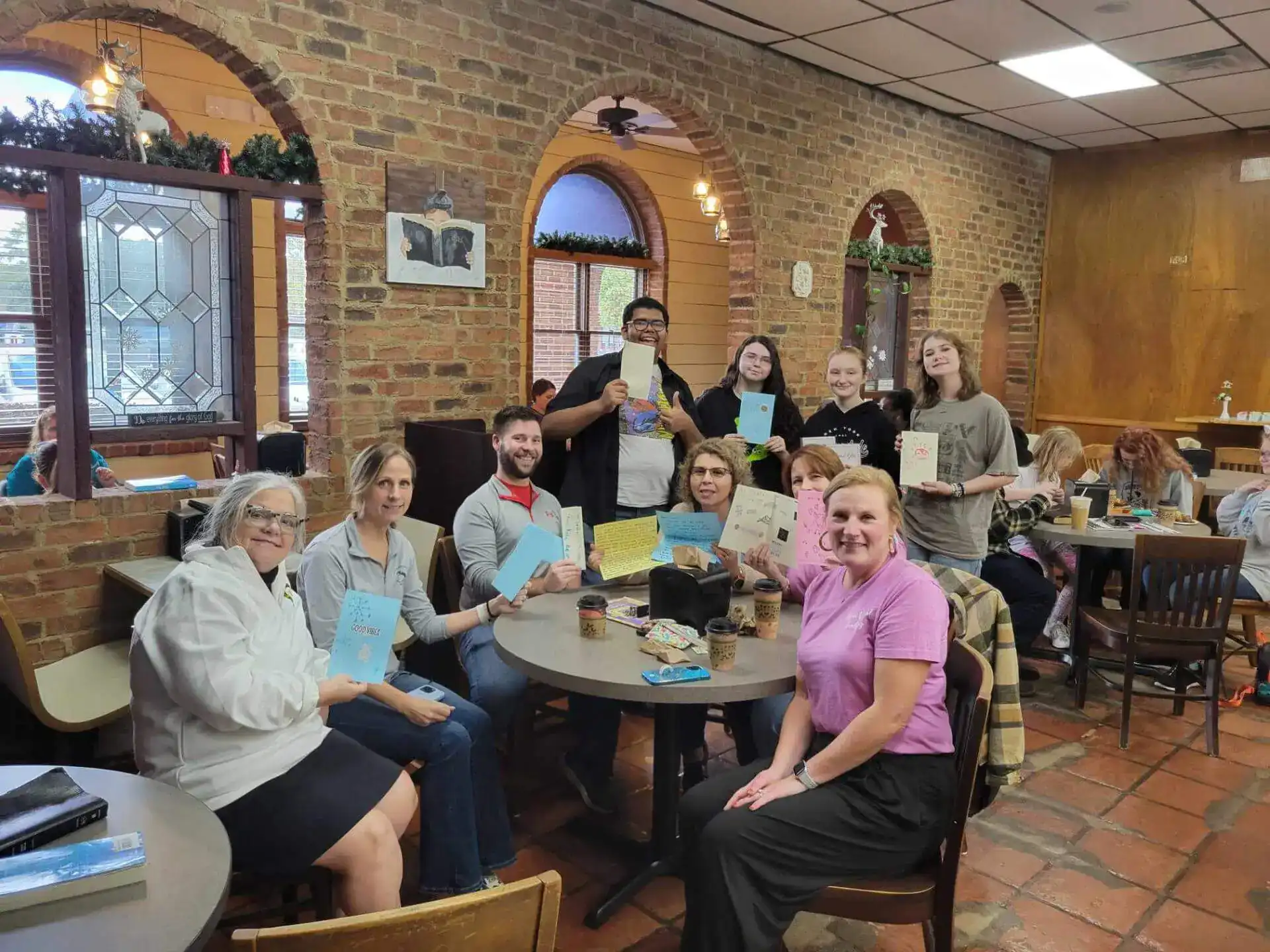 Un grupo de personas sentadas y de pie en una acogedora cafetería con paredes de ladrillo, sosteniendo tarjetas de felicitación hechas a mano.