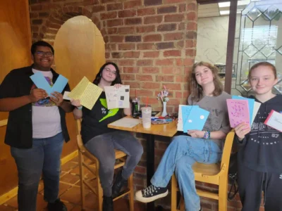Cuatro personas sentadas en un acogedor café, sonriendo y sosteniendo tarjetas hechas a mano.
