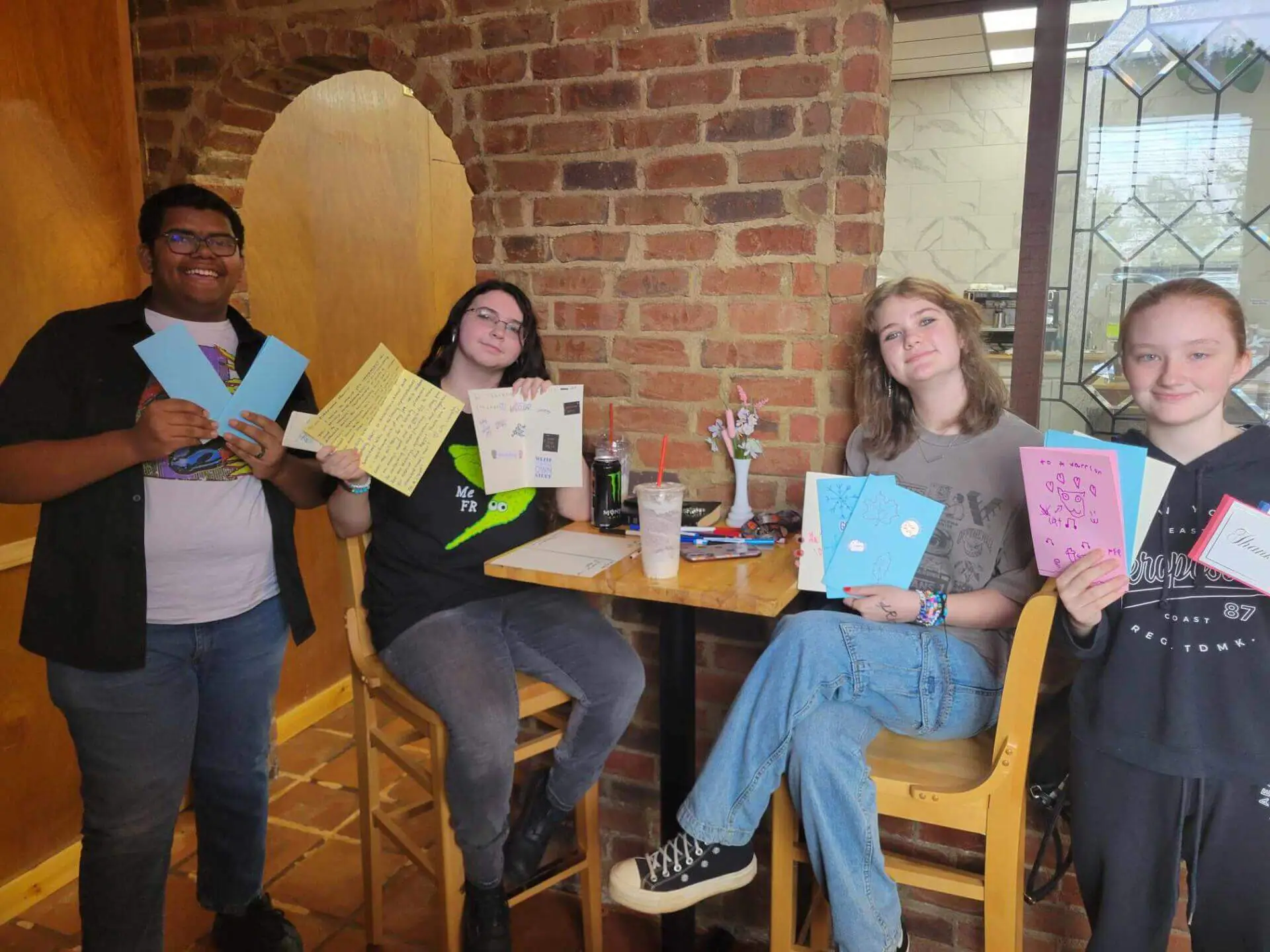 Cuatro personas sentadas en un acogedor café, sonriendo y sosteniendo tarjetas hechas a mano.