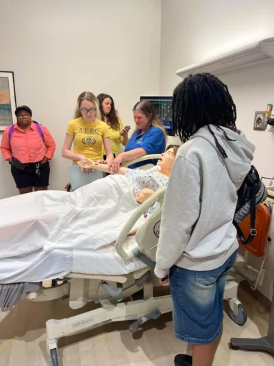 Un grupo de estudiantes en una sala de formación médica observa un maniquí médico en una cama de hospital.