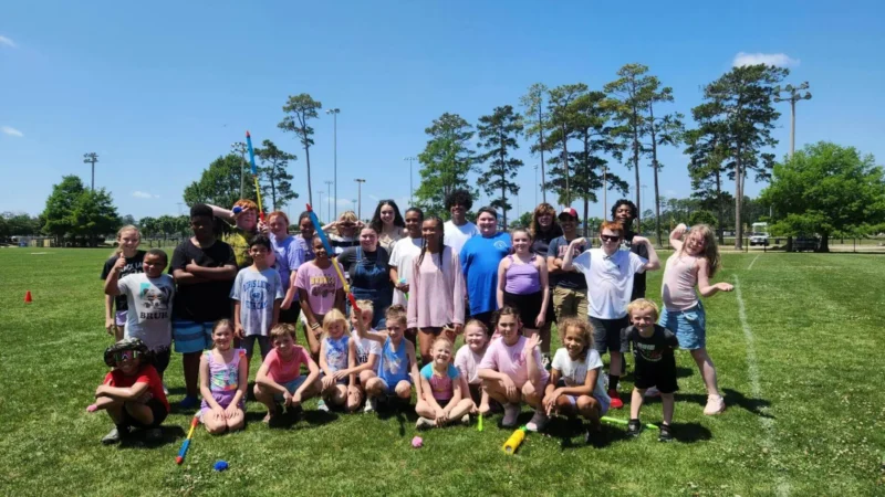 Un grupo de niños y adultos sonrientes posa en un día soleado en un parque, con hierba verde y altos árboles de fondo, transmitiendo alegría y camaradería.