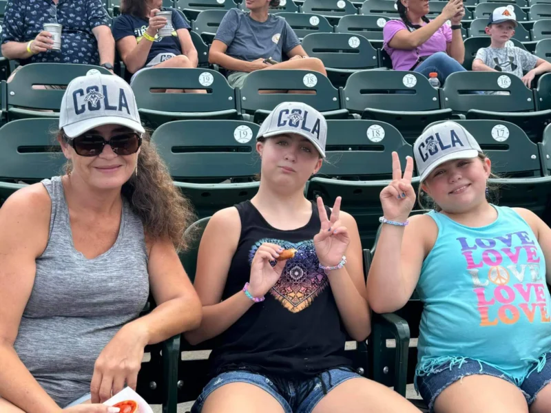 Tres personas con gorras "COLA" sentadas en un estadio.