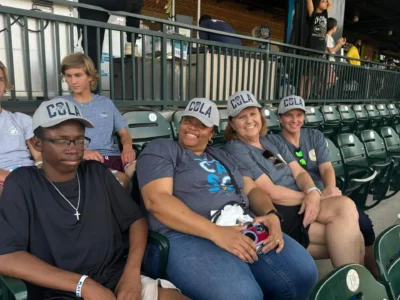 Un grupo de cuatro personas se sienta en los asientos del estadio, sonrientes y con gorras "COLA" y camisetas azules a juego.