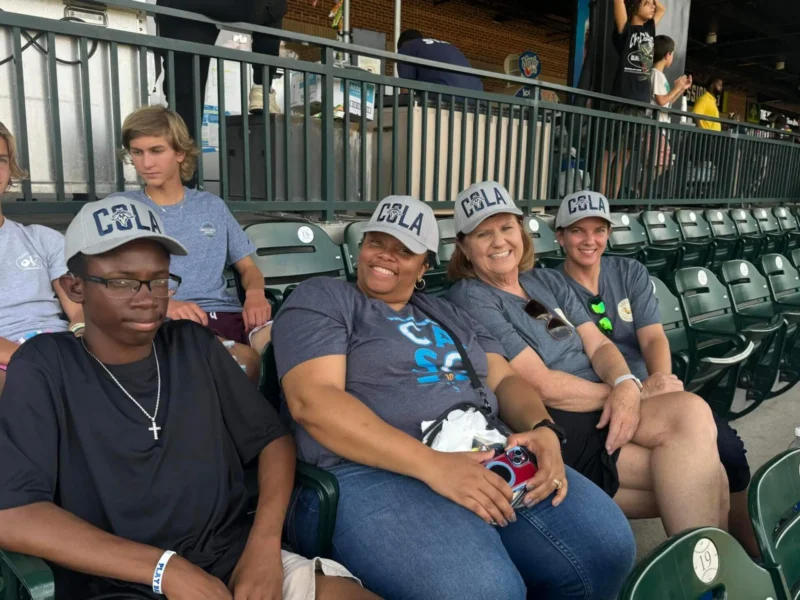 Un grupo de cuatro personas se sienta en los asientos del estadio, sonrientes y con gorras "COLA" y camisetas azules a juego.