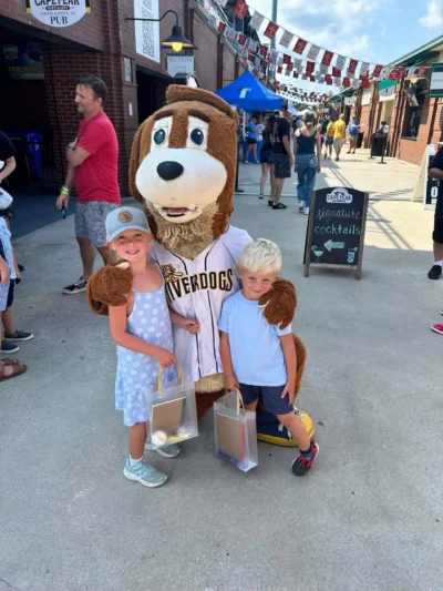 Dos niños sonrientes posan con una simpática mascota canina vestida con una camiseta de béisbol.