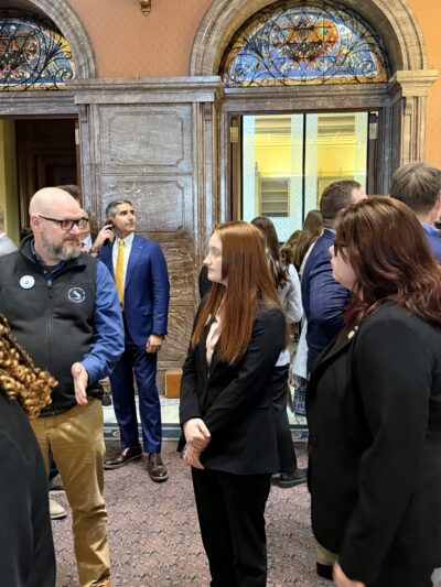 Un grupo de personas vestidas de gala se encuentra reunido en una sala con vitrales ornamentados.